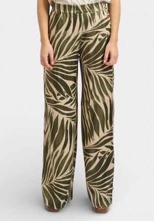 Donna che indossa pantaloni larghi a vita alta con un motivo a grandi foglie in verde oliva e beige, in piedi con le braccia rilassate ai lati.