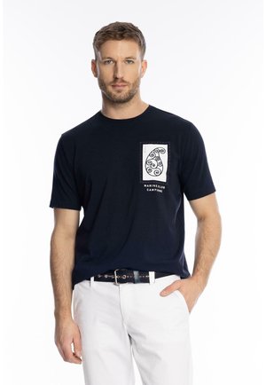 Mann mit kurzem Haar, der ein schwarzes T-Shirt mit Grafik und weiße Hosen trägt, steht mit einer Hand in der Tasche vor weißem Hintergrund.