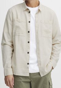 Beige Hemd mit Knöpfen und zwei Brusttaschen, schwarzen Knöpfen und leichtem Gewebe, kombiniert mit einem weißen T-Shirt und grünen Hosen.