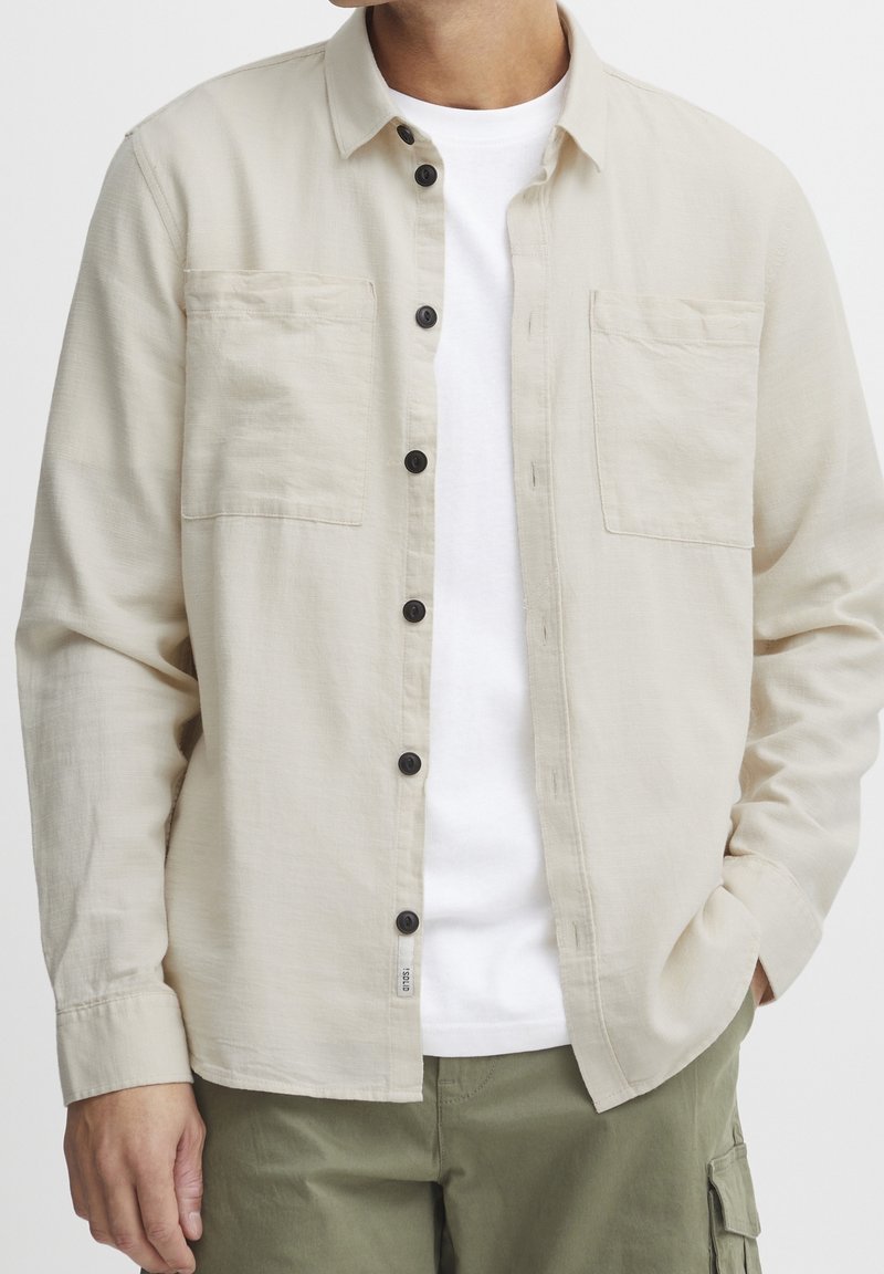 Beige Hemd mit Knöpfen und zwei Brusttaschen, schwarzen Knöpfen und leichtem Gewebe, kombiniert mit einem weißen T-Shirt und grünen Hosen.