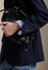 Henkilö, joka käyttää tummansinistä bleiseriä ja vaaleansinistä paitaa, pitää mustaa nahkalaukkua kultasulkijalla, yhdistettynä sinisiin farkkuihin.