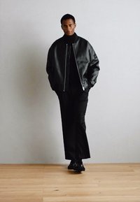 Sort skinnjakke med sølvglidelås, kombinert med en svart turtleneck og løstsittende bukser, mot en lys bakgrunn.