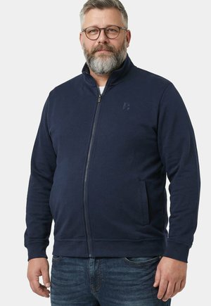 Skægget mand iført briller, en navy lynlåstrøje med sidelommer og blå jeans, står foran en ensfarvet baggrund.