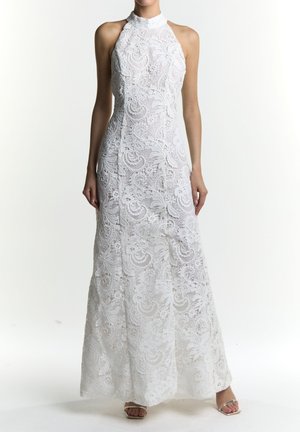 Hvid maxi-kjole i blonder med halsudskæring, der har indviklede blomstermønstre og en flydende silhuet. Sandalerne er sølvfarvede.