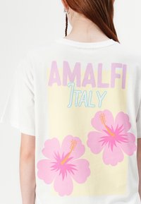 Balta kokvilnas T-krekls ar grafisku dizainu, kurā redzami teksts "AMALFI" un "Itālija" pasteļkrāsās, kā arī divas lielas rozā hibiska ziedi.