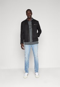 Chaqueta de ante sintético negra con cremallera frontal, camiseta gráfica gris, vaqueros de mezclilla azul claro y zapatillas blancas. Diseño sencillo, acentos mínimos.