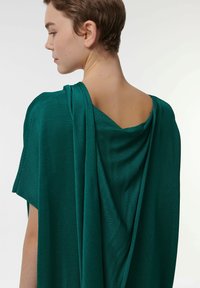 Prenda de punto ligera y verde con un diseño drapeado, que presenta una espalda en V profunda y mangas cortas, texturizada para mayor interés visual.