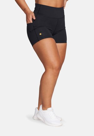 Kobieta ubrana w czarne sportowe szorty z wysokim stanem i bocznymi kieszeniami oraz białe buty do biegania, stojąca bokiem.