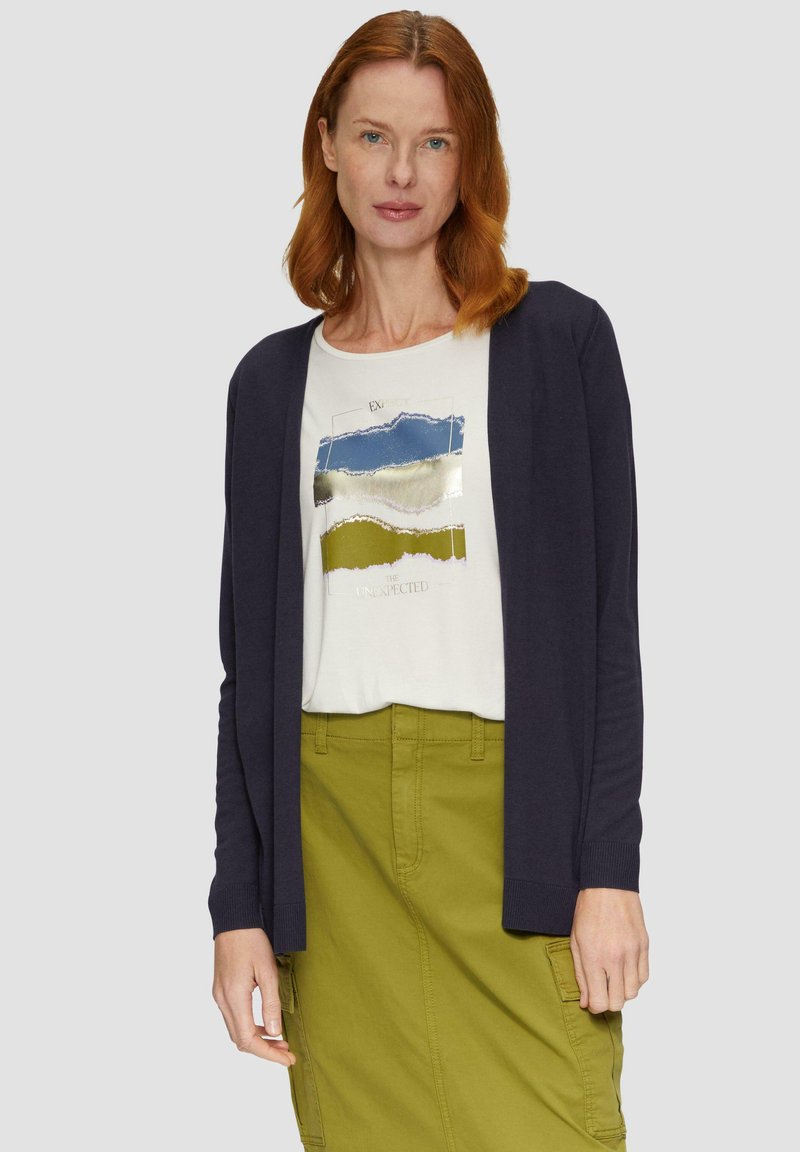 Cardigan bleu marine à manches longues, porté sur une chemise claire avec un motif abstrait multicolore, associé à une jupe verte. Tissu lisse.