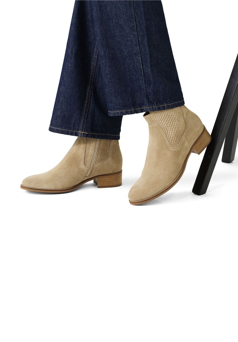 Bottes chelsea en daim beige avec panneaux latéraux texturés et talons carrés en bois, portées avec un pantalon en denim bleu foncé, pieds reposant près des pieds d'une chaise noire.