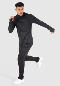 Veste de sport noire avec un col haut et une fermeture éclair, assortie à un pantalon. Les deux présentent une texture lisse et des détails de design minimaux.