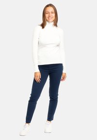Maglione bianco a collo alto con maniche lunghe, abbinato a pantaloni slim-fit blu navy. Il modello indossa sneakers bianche, creando un look informale.