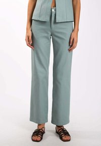 Pantalons larges vert clair en tissu doux, dotés d'une taille haute et d'une fermeture à bouton sur le devant. Associés à des sandales noires à lanières.