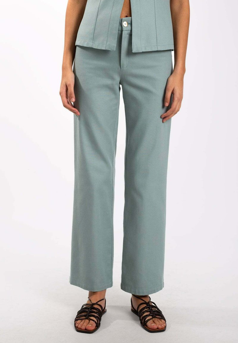 Pantalons larges vert clair en tissu doux, dotés d'une taille haute et d'une fermeture à bouton sur le devant. Associés à des sandales noires à lanières.