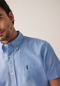 Šviesiai mėlynas trumpomis rankovėmis marškinėliai su mygtukais iš minkštos medžiagos su subtilia tekstūra, su nedideliu siuvinėtu logotipu ant krūtinės ir mygtukų apykakle.