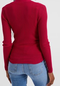 Henkilö, joka on pukeutunut istuvaan pitkähiharaiseen punaisestä ribbikankaasta valmistettuun neuleeseen ja vaaleansinisistä farkuista, näkyy takaa yksinkertaista taustaa vasten.