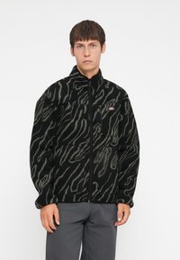 Black fleece jacket with gray zebra stripes, full zip front, high collar, and zippered chest pocket. Soft texture and relaxed fit.