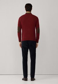 Maglione bordeaux con colletto verde e toppe marroni sui gomiti, abbinato a pantaloni blu navy. Texture liscia, silhouette aderente.