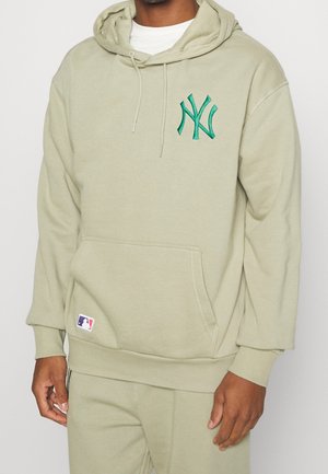 Mężczyzna w jasnobeżowej bluzie z kapturem z zielonym logo New York Yankees i naszywką z logo MLB przy przedniej kieszeni, w zestawie z dopasowanymi spodniami.