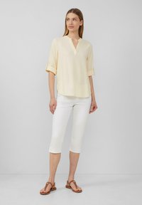 Chemise jaune clair à manches courtes avec un décolleté en V et des poignets roulés, associée à un pantalon blanc court et des sandales plates marron.