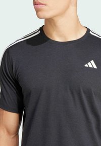 Camiseta deportiva negra de manga corta, con acentos en tres franjas blancas en los hombros y un logo blanco en la esquina inferior derecha.