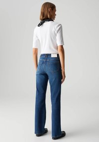 Jeans flare in denim blu con una patch sul retro, abbinati a una maglietta bianca a maniche corte e a una sciarpa nera al collo. Tessuto morbido e vestibilità casual.