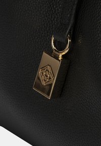 Zwarte leren handtas met een textuur, voorzien van een gouden metalen charm met een geometrisch logo dat aan een ring is bevestigd.