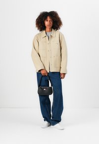 Beige utilityjakke med to frontlommer, blå vide jeans, hvide sneakers og en lille sort håndtaske med et farvet logo.