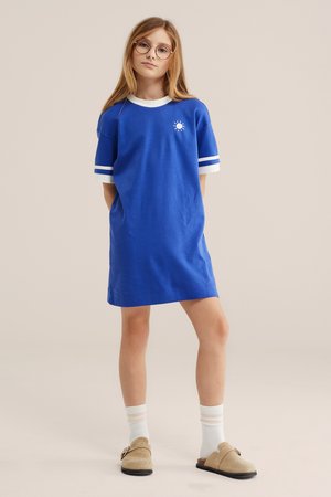 Mlajša deklica z dolgimi blond lasmi in očali, oblečena v modro krilo s kratkimi rokavi in belim robom ter bele nogavice in bež čevlje brez vezalk.