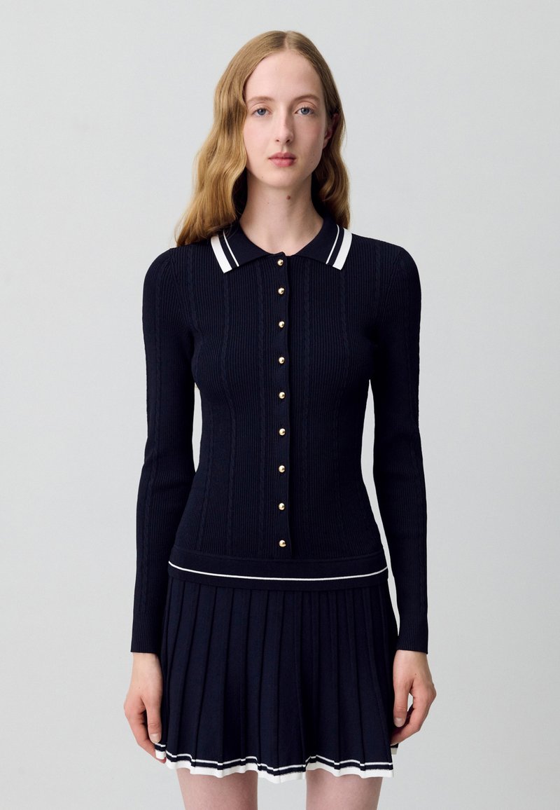 Giovane donna con lunghi capelli mosso che indossa un maglione blu navy aderente con bottoni e finiture bianche, abbinato a una gonna plissettata.