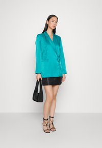 Blazer en satin turquoise avec taille cintrée, doté d'un col à revers, de poches latérales et de manches longues, associé à une mini jupe en cuir noire.