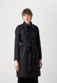 Manteau noir à double boutonnage, quincaillerie en argent et ceinture assortie. Présente des accents gris contrastants sur les poches et le col.