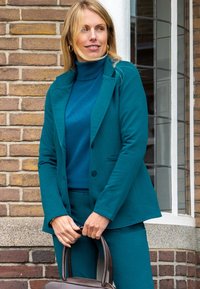 Teal blazer met gekartelde revers, twee zakken en knopen. Draagt het over een bijpassende coltrui, gecombineerd met teal broek en houdt een bruine handtas vast.