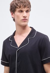 Μαύρο πιτζάμα πουκάμισο με κοντά μανίκια, γιακά με εγκοπή και λευκή λεπτομέρεια με ρέλι. Διαθέτει ένα κουμπί μπροστά για κλείσιμο.