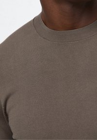 Närbild av en person som bär en enkel brun sweatshirt, där man ser halsringningen och övre delen av bröstet.