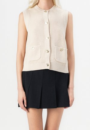 Femme portant un gilet en maille beige sans manches boutonné avec des poches avant et une jupe plissée noire.