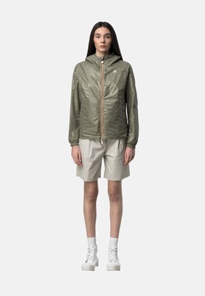 Donna con capelli lunghi e scuri che indossa una giacca con cappuccio verde oliva, pantaloncini beige fino al ginocchio, calzini bianchi e stivali bianchi con lacci.