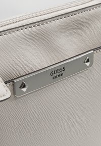 Primer plano de una tela de bolsa gris texturizada con una placa de metal plateada grabada "GUESS EST. 1981" sujeta por dos tachuelas en forma de pirámide.
