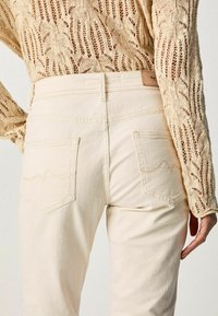 Jean beige clair à taille haute, avec des poches arrière visibles et des détails de couture subtils. Porté avec un pull en maille beige texturé.