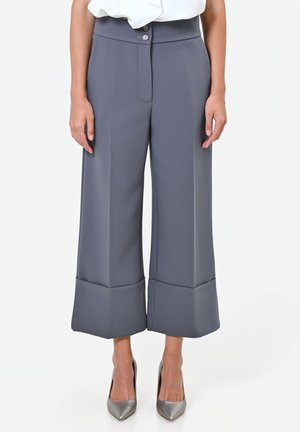 Vrouw draagt een grijze, hoog getailleerde, wijd uitlopende cropped broek met opgerolde pijpen en een tailleband met twee knopen, gecombineerd met zilverkleurige pumps met puntige neus.