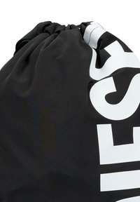 Czarny materiałowy worek ze sznurkiem, ozdobiony białym napisem, z marszczonym otworem i teksturowaną powierzchnią.