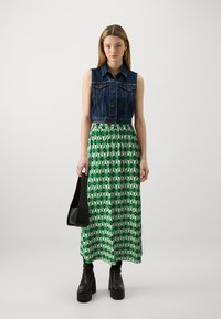 Gilet en denim, jupe maxi verte à motifs géométriques, sac noir et bottes à plateforme. Tissu lisse, taille ajustée et silhouette ample.