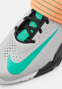 Zapatilla deportiva gris con cordones negros, swoosh turquesa y parte superior texturizada en gris. Presenta una correa naranja para mayor soporte.