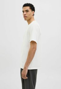 Tricou scurt cu mânecă scurtă, de culoare crem deschis, cu guler rotund și croi lejer, purtat cu pantaloni gri închis. Prezintă tatuaje vizibile pe braț.