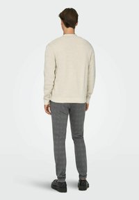 Homme debout de dos, portant un pull beige clair, un pantalon gris ajusté et des chaussures de ville noires sur un fond uni.