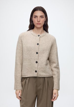 Giovane donna con lunghi capelli scuri che indossa un maglione di lana beige con bottoni e pantaloni marroni larghi, in piedi davanti a uno sfondo chiaro e semplice.