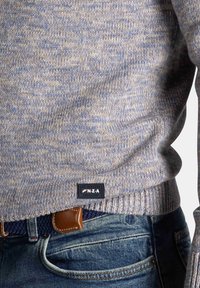 Lichtblauwe gebreide trui met een gestructureerde afwerking, voorzien van een zwart logo-label aan de zoom, gecombineerd met blauwe spijkerbroek en een bruine riem.