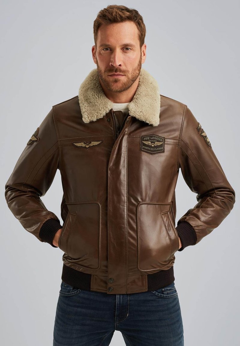 Giacca bomber in pelle marrone con colletto in pelliccia sintetica, dotata di due tasche frontali, toppe ricamate e polsini e orlo a coste.
