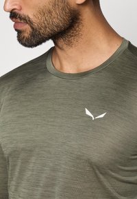 Olivnozelena majica s kratkimi rokavi in okroglim izrezom z gladko teksturo; na levi strani prsnega koša je majhna bela oznaka.