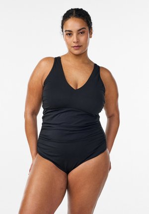 Kobieta o kręconych włosach, ubrana w czarny jednoczęściowy kostium kąpielowy, stojąca pewnie na jednolitym białym tle.